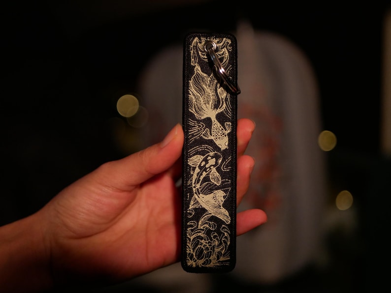 K&ouml;nnte beinhalten: Schwarzes rechteckiges Etui mit einem goldenen Koi-Fisch-Design. Das Etui hat einen Metallring zur Befestigung. Die Koi-Fische schwimmen in einem detaillierten, flie&szlig;enden Muster. Der Hintergrund ist dunkel, mit verschwommenen Lichtern.