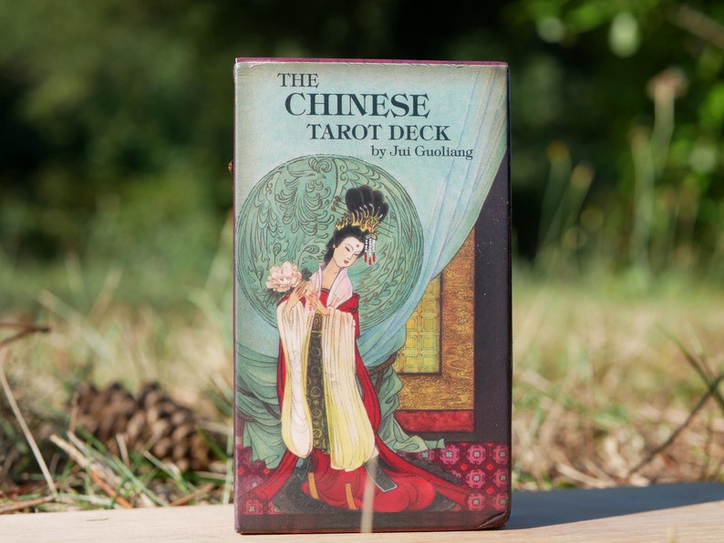 May include: A tarot deck box titled "THE CHINESE TAROT DECK" by Jui Guoliang. The box features an illustration of a woman in traditional Chinese attire, holding flowers. The background is a mix of green, blue, and gold.