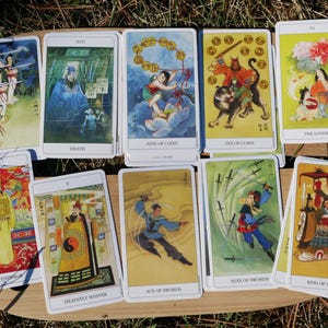 May include: A collection of tarot cards with Asian-inspired illustrations. The cards feature images of figures, symbols, and scenes, including "Death," "Nine of Coins," and "The Lovers." The cards are arranged on a wooden surface.