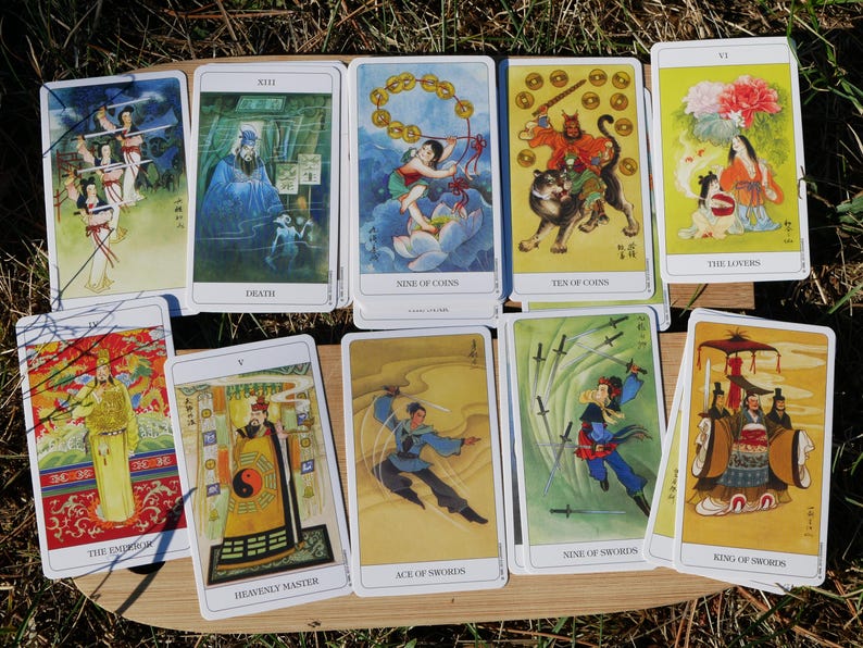 May include: A collection of tarot cards with Asian-inspired illustrations. The cards feature images of figures, symbols, and scenes, including "Death," "Nine of Coins," and "The Lovers." The cards are arranged on a wooden surface.