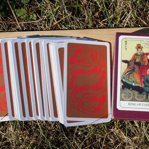 May include: A deck of tarot cards with a burgundy box. The card backs are brown with a red swirl design. The box shows a card with an illustration of a robed figure and the text "KING OF COINS".