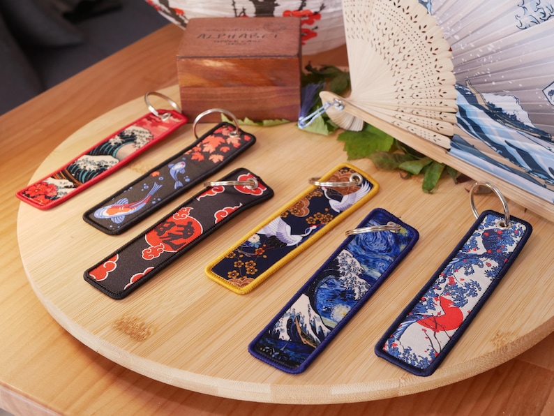 K&ouml;nnte beinhalten: Eine Sammlung von Schl&uuml;sselanh&auml;ngern im japanischen Stil mit verschiedenen Designs, darunter Wellen, Koi-Fische und Kraniche. Jeder Schl&uuml;sselanh&auml;nger ist rechteckig mit einem Metallring und einem farbenfrohen Stoffdesign. Sie sind auf einem Holztablett angeordnet.