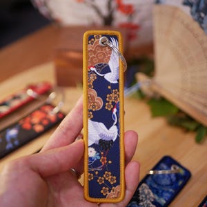 K&ouml;nnte beinhalten: Ein rechteckiger Schl&uuml;sselanh&auml;nger mit japanisch inspiriertem Design. Der marineblaue Stoff zeigt wei&szlig;e Kraniche, goldene Blumen und braune Wolken. Die Kanten sind mit goldenen N&auml;hten versehen. Oben ist ein silberner Schl&uuml;sselring befestigt. Weitere Schl&uuml;sselanh&auml;nger mit &auml;hnlichen Designs sind sichtbar.