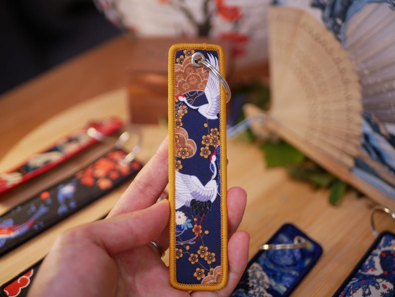 K&ouml;nnte beinhalten: Ein rechteckiger Schl&uuml;sselanh&auml;nger mit japanisch inspiriertem Design. Der marineblaue Stoff zeigt wei&szlig;e Kraniche, goldene Blumen und braune Wolken. Die Kanten sind mit goldenen N&auml;hten versehen. Oben ist ein silberner Schl&uuml;sselring befestigt. Weitere Schl&uuml;sselanh&auml;nger mit &auml;hnlichen Designs sind sichtbar.