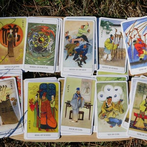 May include: A collection of tarot cards featuring colorful illustrations of figures in traditional Asian attire. The cards are arranged on a wooden tray, with titles such as "Wheel of Fortune" and "Seven of Swords" visible.