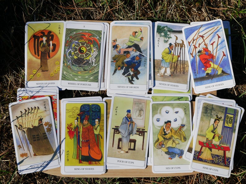 May include: A collection of tarot cards featuring colorful illustrations of figures in traditional Asian attire. The cards are arranged on a wooden tray, with titles such as "Wheel of Fortune" and "Seven of Swords" visible.