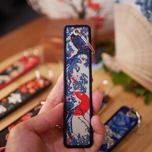 K&ouml;nnte beinhalten: Ein rechteckiger Schl&uuml;sselanh&auml;nger mit japanisch inspiriertem Design mit roter Sonne, blauen Wellen und wei&szlig;en Akzenten. Der Schl&uuml;sselanh&auml;nger hat einen marineblauen Rand und einen silbernen Schl&uuml;sselring. Weitere Schl&uuml;sselanh&auml;nger mit &auml;hnlichen Designs sind sichtbar.