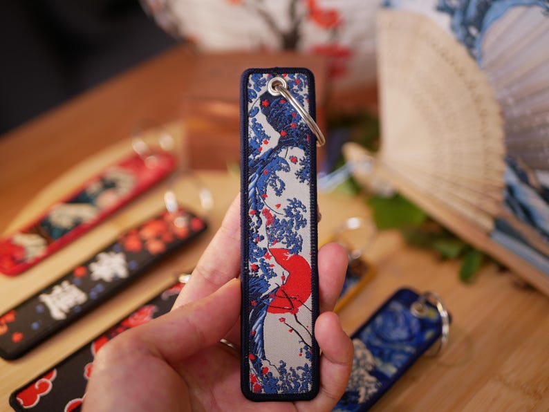 K&ouml;nnte beinhalten: Ein rechteckiger Schl&uuml;sselanh&auml;nger mit japanisch inspiriertem Design mit roter Sonne, blauen Wellen und wei&szlig;en Akzenten. Der Schl&uuml;sselanh&auml;nger hat einen marineblauen Rand und einen silbernen Schl&uuml;sselring. Weitere Schl&uuml;sselanh&auml;nger mit &auml;hnlichen Designs sind sichtbar.
