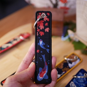 K&ouml;nnte beinhalten: Schwarzer rechteckiger Schl&uuml;sselanh&auml;nger mit einem silbernen Ring. Der Schl&uuml;sselanh&auml;nger zeigt ein Design aus roten Ahornbl&auml;ttern und blau-roten Koi-Fischen. Der Hintergrund ist schwarz mit einem roten Rand.