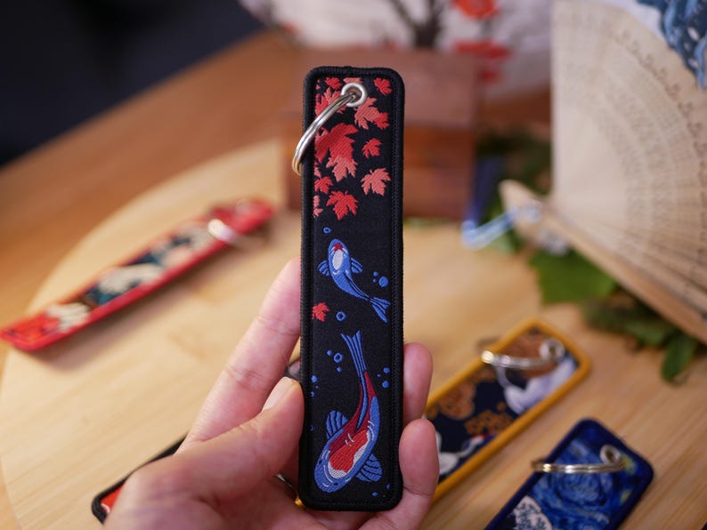 K&ouml;nnte beinhalten: Schwarzer rechteckiger Schl&uuml;sselanh&auml;nger mit einem silbernen Ring. Der Schl&uuml;sselanh&auml;nger zeigt ein Design aus roten Ahornbl&auml;ttern und blau-roten Koi-Fischen. Der Hintergrund ist schwarz mit einem roten Rand.