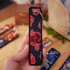 K&ouml;nnte beinhalten: Schwarzer rechteckiger Schl&uuml;sselanh&auml;nger mit rotem Wolken- und Kreismuster. Der Schl&uuml;sselanh&auml;nger hat einen Metallring und einen schwarzen Rand. Das Design enth&auml;lt japanische Schriftzeichen. Weitere Schl&uuml;sselanh&auml;nger mit &auml;hnlichen Designs sind sichtbar.