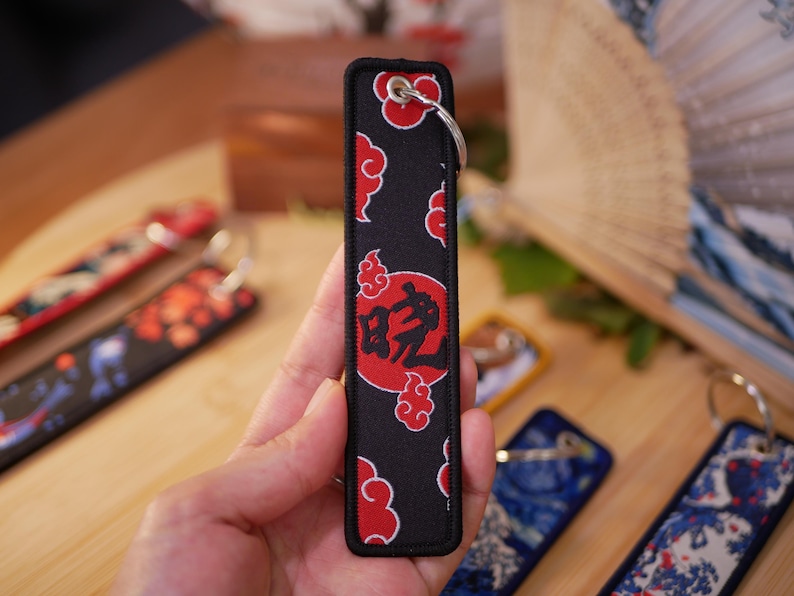 K&ouml;nnte beinhalten: Schwarzer rechteckiger Schl&uuml;sselanh&auml;nger mit rotem Wolken- und Kreismuster. Der Schl&uuml;sselanh&auml;nger hat einen Metallring und einen schwarzen Rand. Das Design enth&auml;lt japanische Schriftzeichen. Weitere Schl&uuml;sselanh&auml;nger mit &auml;hnlichen Designs sind sichtbar.