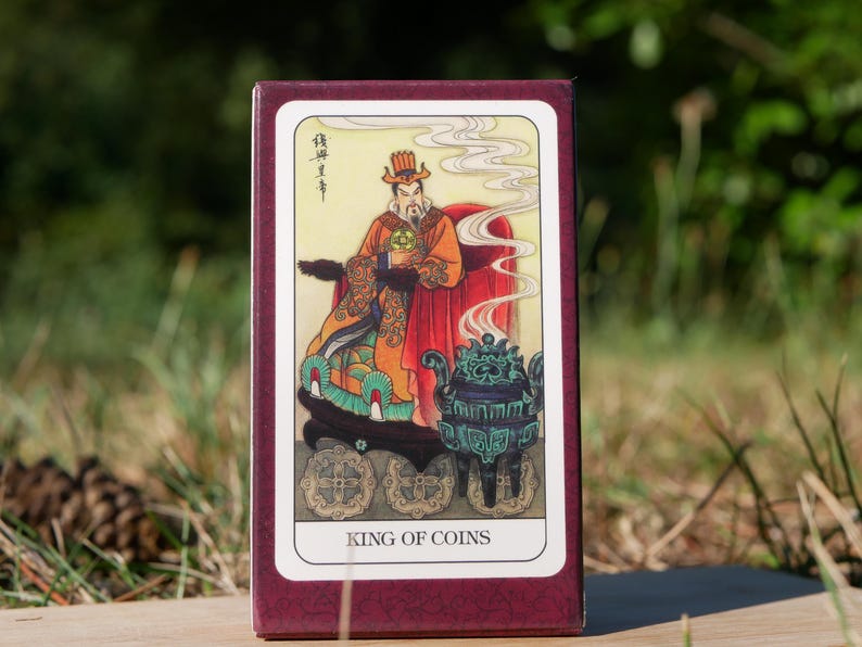 May include: A tarot card box featuring the King of Coins. The illustration depicts a robed figure with a beard, holding a coin, and seated on a throne. The card has a burgundy border and the words "KING OF COINS" are printed at the bottom.