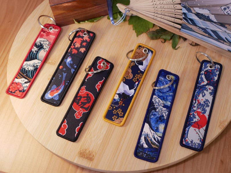 K&ouml;nnte beinhalten: Bestickte Schl&uuml;sselanh&auml;nger mit japanisch inspirierten Designs. Die Schl&uuml;sselanh&auml;nger zeigen verschiedene Muster, darunter Wellen, Koi-Fische, Kraniche und florale Motive. Jeder Schl&uuml;sselanh&auml;nger hat einen Metallring zur Befestigung und ist rechteckig.