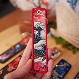 K&ouml;nnte beinhalten: Ein rechteckiger Schl&uuml;sselanh&auml;nger mit japanischem Wellenmuster und rotem Rand. Das Design zeigt eine gro&szlig;e Meereswelle in Blau und Wei&szlig;, mit einem roten Blumenmuster am unteren Rand. Ein silberner Schl&uuml;sselring ist befestigt.