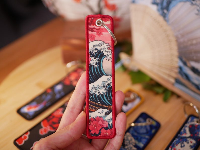 K&ouml;nnte beinhalten: Ein rechteckiger Schl&uuml;sselanh&auml;nger mit japanischem Wellenmuster und rotem Rand. Das Design zeigt eine gro&szlig;e Meereswelle in Blau und Wei&szlig;, mit einem roten Blumenmuster am unteren Rand. Ein silberner Schl&uuml;sselring ist befestigt.