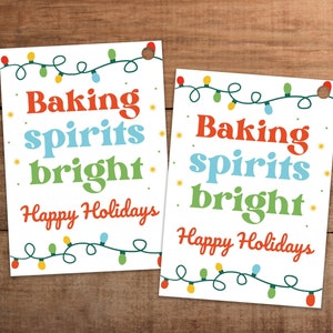 Puede incluir: Dos tarjetas rectangulares blancas con el texto "Baking spirits bright" en rojo, azul y verde, y "Happy Holidays" en rojo. Las tarjetas están decoradas con una guirnalda de luces navideñas de colores y pequeñas estrellas amarillas.