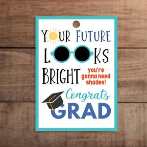 May include: A rectangular white gift tag with a teal border and a hole at the top. It features a yellow sun, black sunglasses, a black graduation cap, and the text: Your future looks bright, you're gonna need shades! Congrats GRAD.