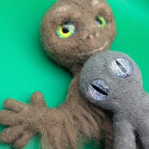 May include: Handmade felted alien dolls in brown and gray. The larger doll has large eyes with yellow, green, and blue irises. The smaller doll has gray eyes with a white and black pattern. The dolls are set against a green background.