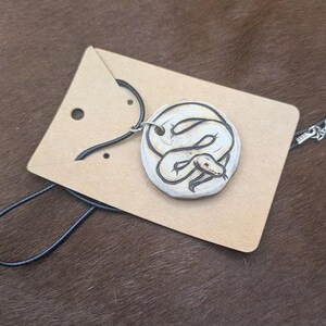 Può includere: Una collana con ciondolo in legno e un cordoncino nero. Il ciondolo è un pezzo di legno rotondo con un'illustrazione in bianco e nero di un serpente.