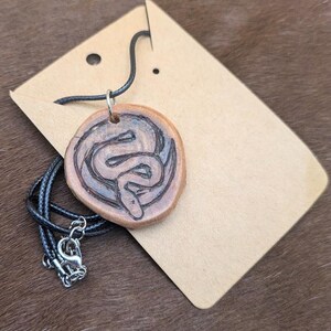 Può includere: Una collana con un ciondolo in legno e un cordoncino nero. Il ciondolo è rotondo e presenta un design a serpente intagliato.