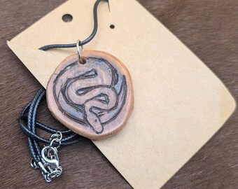 May include: A wooden pendant necklace with a black cord. The pendant is round and features a carved snake design.