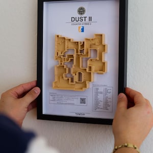 Pode incluir: Obra de arte emoldurada com um mapa 3D bege do mapa Dust II Counter-Strike 2. A obra de arte est&aacute; em uma moldura preta e inclui o logotipo e o texto do jogo. O mapa &eacute; uma representa&ccedil;&atilde;o arquitet&ocirc;nica detalhada.