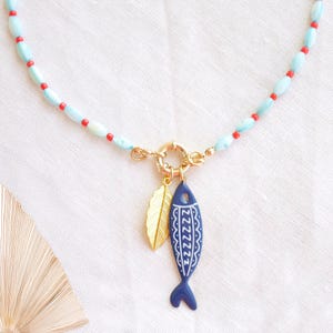 Può includere: Una collana con perline ovali azzurre e piccole perline rotonde rosse. Una chiusura circolare dorata collega due ciondoli: una foglia dorata e un pesce blu scuro con dettagli decorativi a linee bianche.
