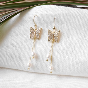 May include: Gold butterfly earrings with dangling pearl accents. The butterflies are adorned with clear rhinestones.