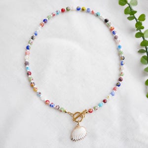 May include: A colourful beaded necklace with a gold clasp and a seashell pendant. The necklace features small, multi-coloured beads and a white seashell pendant with gold trim. The necklace is approximately 40 cm long.