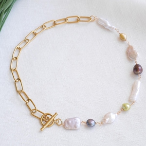 Pode incluir: Um colar dourado com uma combinação de elos de corrente e uma fileira de pérolas de formato irregular em várias cores. As pérolas incluem tons brancos, lavanda, castanhos e verdes. O colar tem um fecho de alternância.