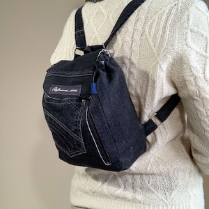 May include: A dark denim backpack with a small pocket and a zipper. The backpack has adjustable straps and a label that reads "Reitmans JEANS". The bag is worn over the shoulder.