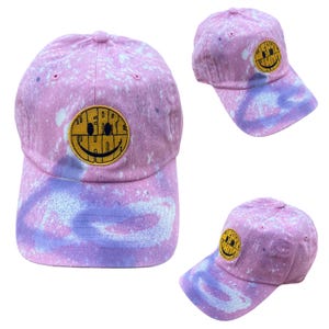 May include: A pink baseball cap with a tie-dye effect, featuring a yellow smiley face patch. The cap has a curved brim and a button on top. The smiley face patch has the word "WEIRD" above the smiley face.