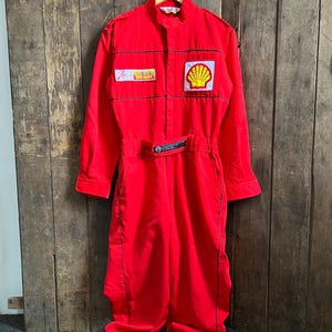 Puede incluir: Mono de carreras rojo brillante con ribetes negros y un parche con el logotipo de Shell. El mono tiene mangas largas, cuello alzado y cinturón. El logotipo de Shell es una concha amarilla sobre fondo rojo. El mono está colgado de una percha de madera.