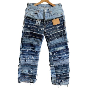 Op de afbeelding: Blauwe spijkerbroek gemaakt van meerdere lagen denimstof, elke laag is gefranst en aan elkaar genaaid. De spijkerbroek heeft een klassieke vijf-zak stijl en een bruin leren patch op de achterkant.