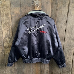 Puede incluir: Una chaqueta bomber de satén negro con cuello alto y forro interior de color claro. La parte trasera muestra el texto bordado en blanco Racing Development y el acrónimo TRD bordado en rojo. Tiene puños y dobladillo elásticos.