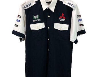 Op de afbeelding: Een donkerblauw, kort mouwig overhemd met witte accenten. Het overhemd heeft twee borstzakken en heeft logo's van Sparco, Mitsubishi Motors en het Wereldkampioenschap Rally.