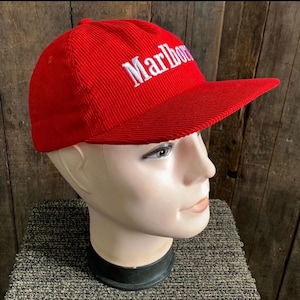 Pode incluir: Boné de beisebol vermelho de veludo cotelê com a palavra "Marlboro" bordada em branco na frente. O boné tem uma aba curva e uma coroa estruturada. O chapéu é exibido em uma cabeça de manequim.