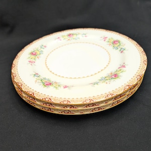 May include: Three stacked cream-colored plates with floral and gold trim. The plates have a central floral design with pink, yellow, and blue flowers. The outer rim features a detailed gold and burgundy pattern.