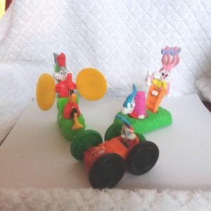 May include: A set of four plastic toy figures of Bugs Bunny and Tweety Bird. The figures are in various poses, including Bugs Bunny driving a red race car, Tweety Bird playing the drums, and Bugs Bunny riding a green slide. The figures are all brightly colored and have a cartoonish style.