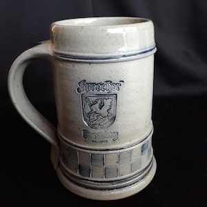 Pode incluir: Uma caneca de cerveja de cerâmica cinza com um design xadrez azul e branco. A caneca apresenta um logotipo da Sprecher Brewing, fundada em 1985.