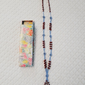 May include: A long blue and brown beaded necklace with a tassel at the bottom. The necklace is in a floral patterned box.