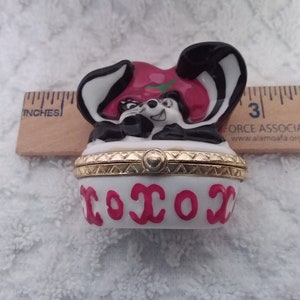 May include: A white porcelain trinket box with a gold trim and a red and black cartoon raccoon design. The lid of the box has the word "XOXO" written in pink.
