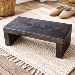 May include: A dark stained wooden step stool with a rectangular top and two supporting legs. The wood grain is visible. The stool is on a beige rug with a fringed edge. A pair of shoes is in the background.