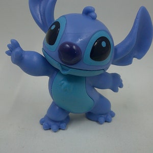 Puede incluir: Una figura de pl&aacute;stico azul de Stitch de la pel&iacute;cula de Disney Lilo & Stitch. Stitch est&aacute; de pie con los brazos extendidos y tiene una gran sonrisa en la cara.