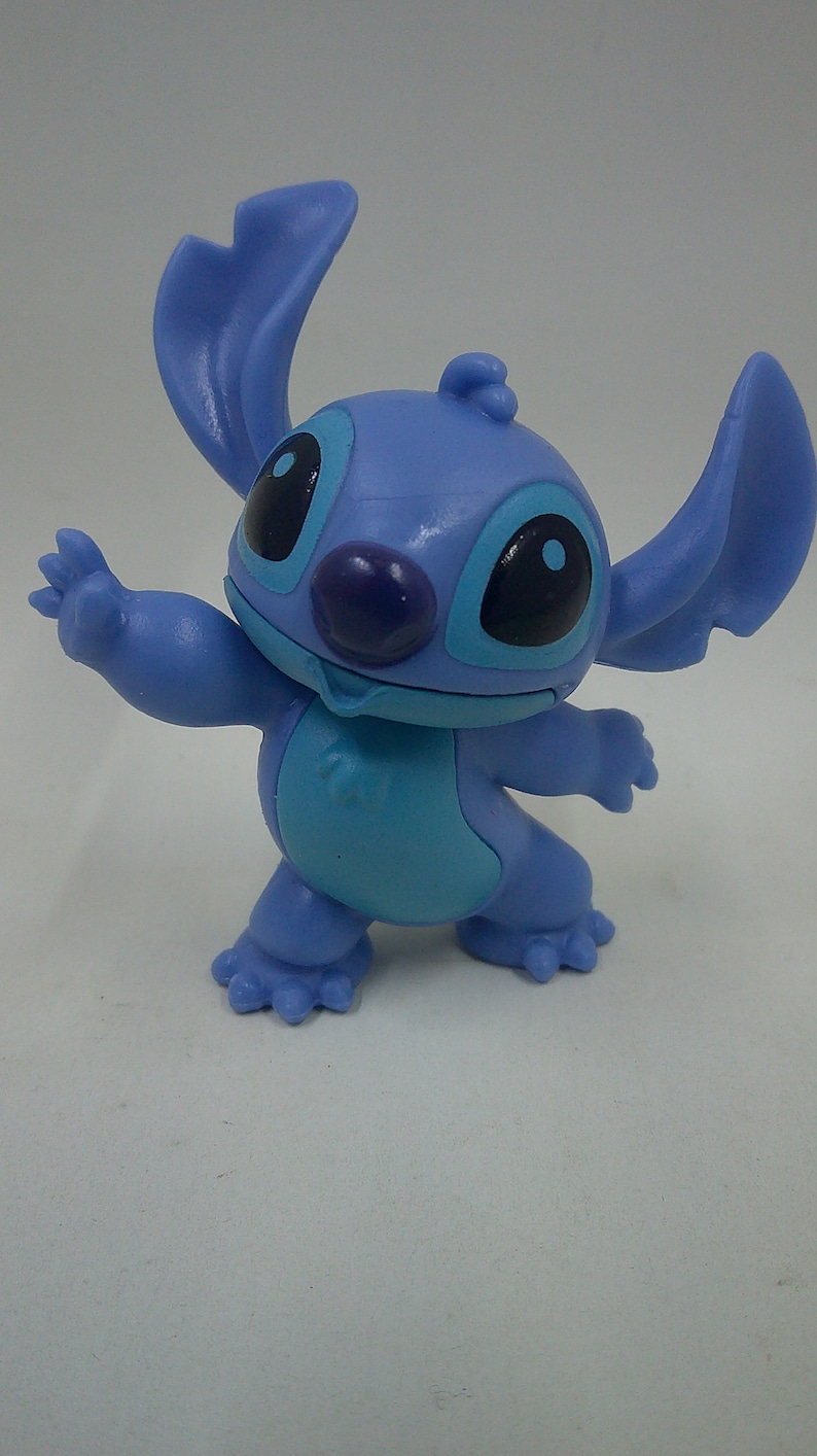 Puede incluir: Una figura de pl&aacute;stico azul de Stitch de la pel&iacute;cula de Disney Lilo & Stitch. Stitch est&aacute; de pie con los brazos extendidos y tiene una gran sonrisa en la cara.