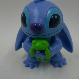 Puede incluir: Una figura azul de Stitch sosteniendo una rana verde. La figura est&aacute; hecha de pl&aacute;stico y tiene un aspecto de dibujos animados.