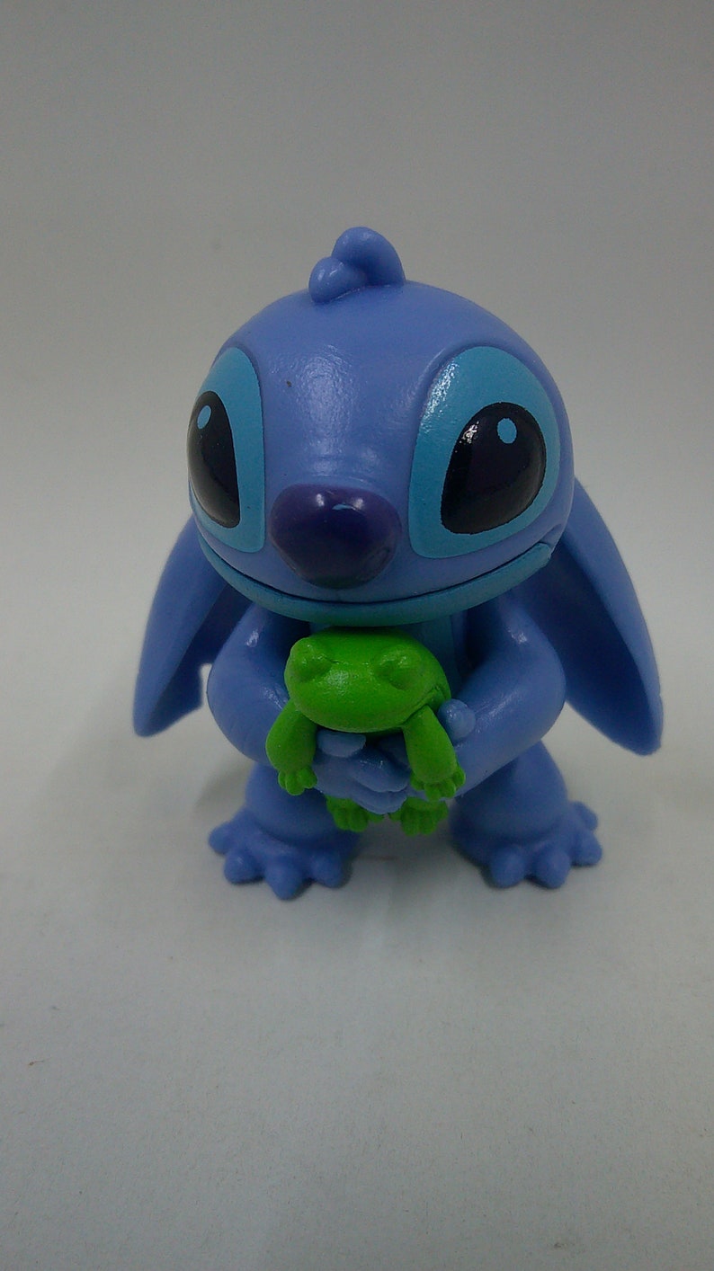 Puede incluir: Una figura azul de Stitch sosteniendo una rana verde. La figura est&aacute; hecha de pl&aacute;stico y tiene un aspecto de dibujos animados.