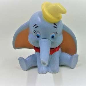 May include: A blue and orange plastic toy elephant figurine with a yellow hat and a red bow tie. The elephant is sitting with its trunk curled up.