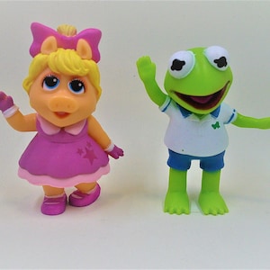 May include: A pink pig figurine with a yellow bow and a green frog figurine wearing a blue shirt and shorts. Both figurines are smiling.